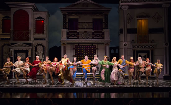 The 17 cast members in costume do a kick-line in front of the three-building cardboard-like set.